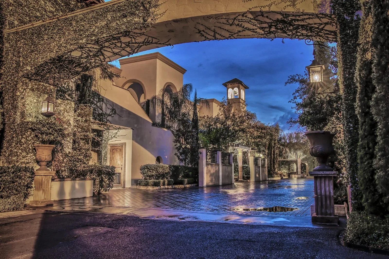 The Bell Tower on 34th entry court at blue hour with an ivy-draped arch, glowing lanterns, and the bell tower beyond, architecture reminiscent of grand Italian villas and haciendas