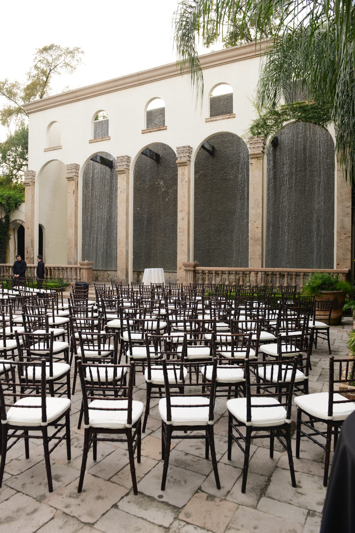 Rows of ceremony chairs set in the daylight courtyard before the arched stone Waterwall