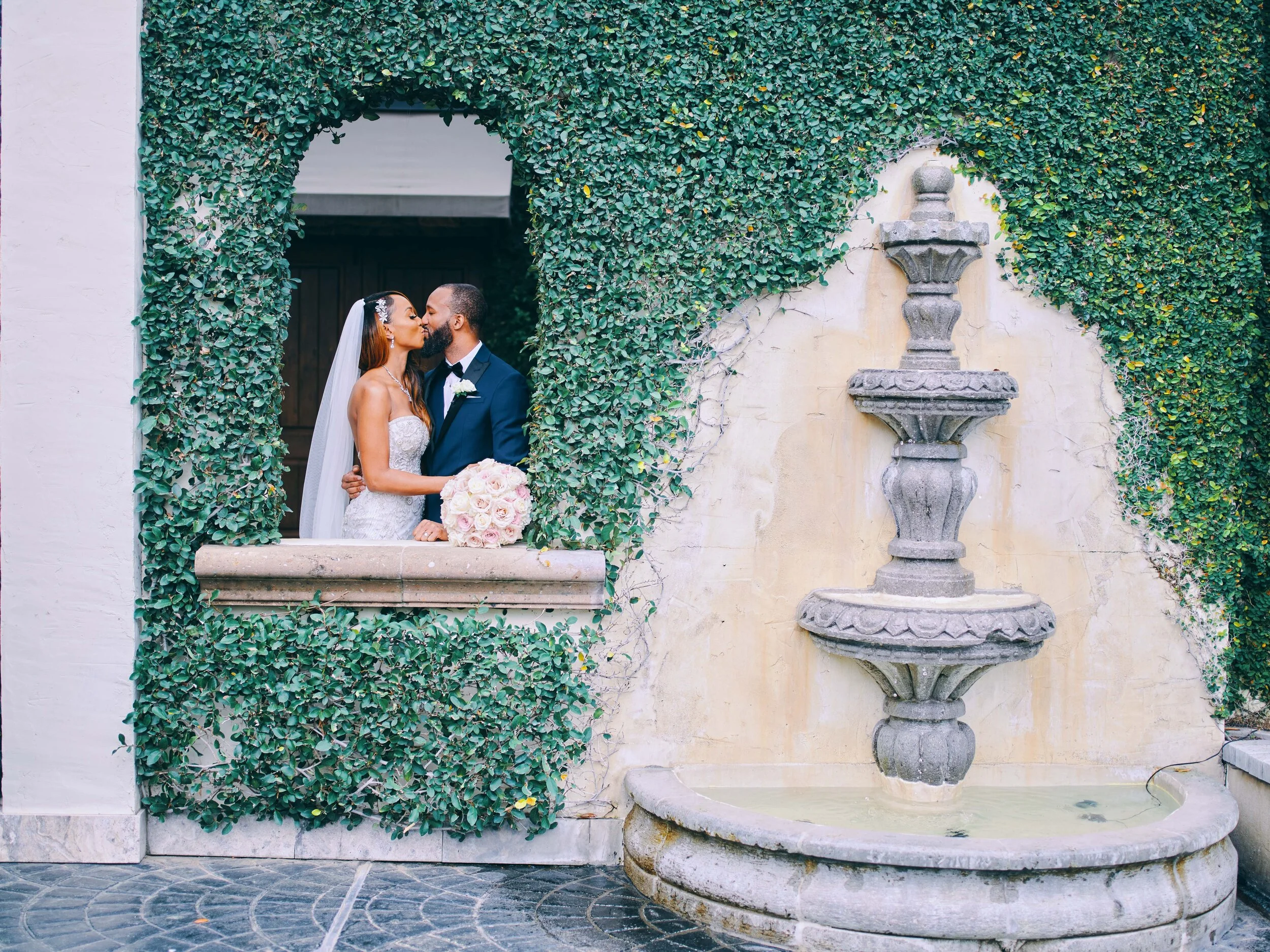 Bride and groom beneath the bright ivy archway by the stone fountain at The Bell Tower on 34th